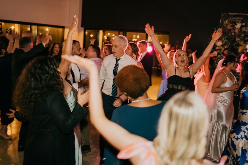 Wedding guests dance with raised hands on a lively dance floor illuminated by pink and amber lighting.