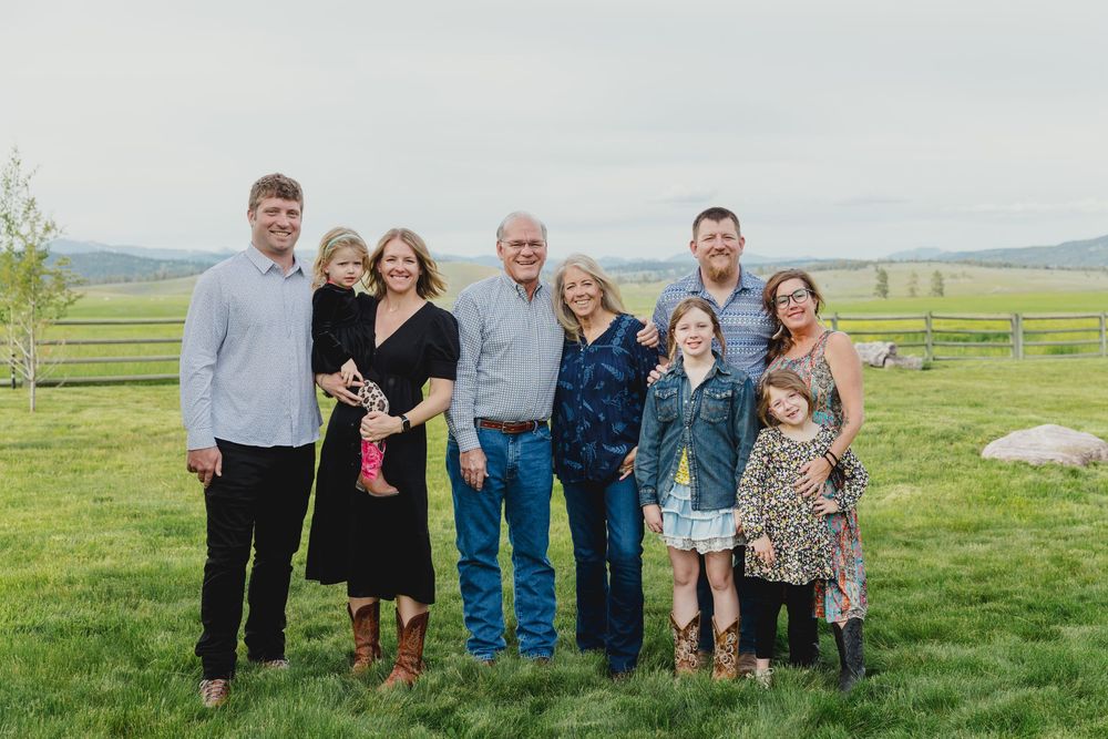 Multi-generational family smiling together outdoors on a green farm field with scenic hills in the background.