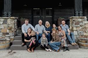 Large family group smiling on rustic stone porch steps with wooden barrels, casual outdoor portrait.