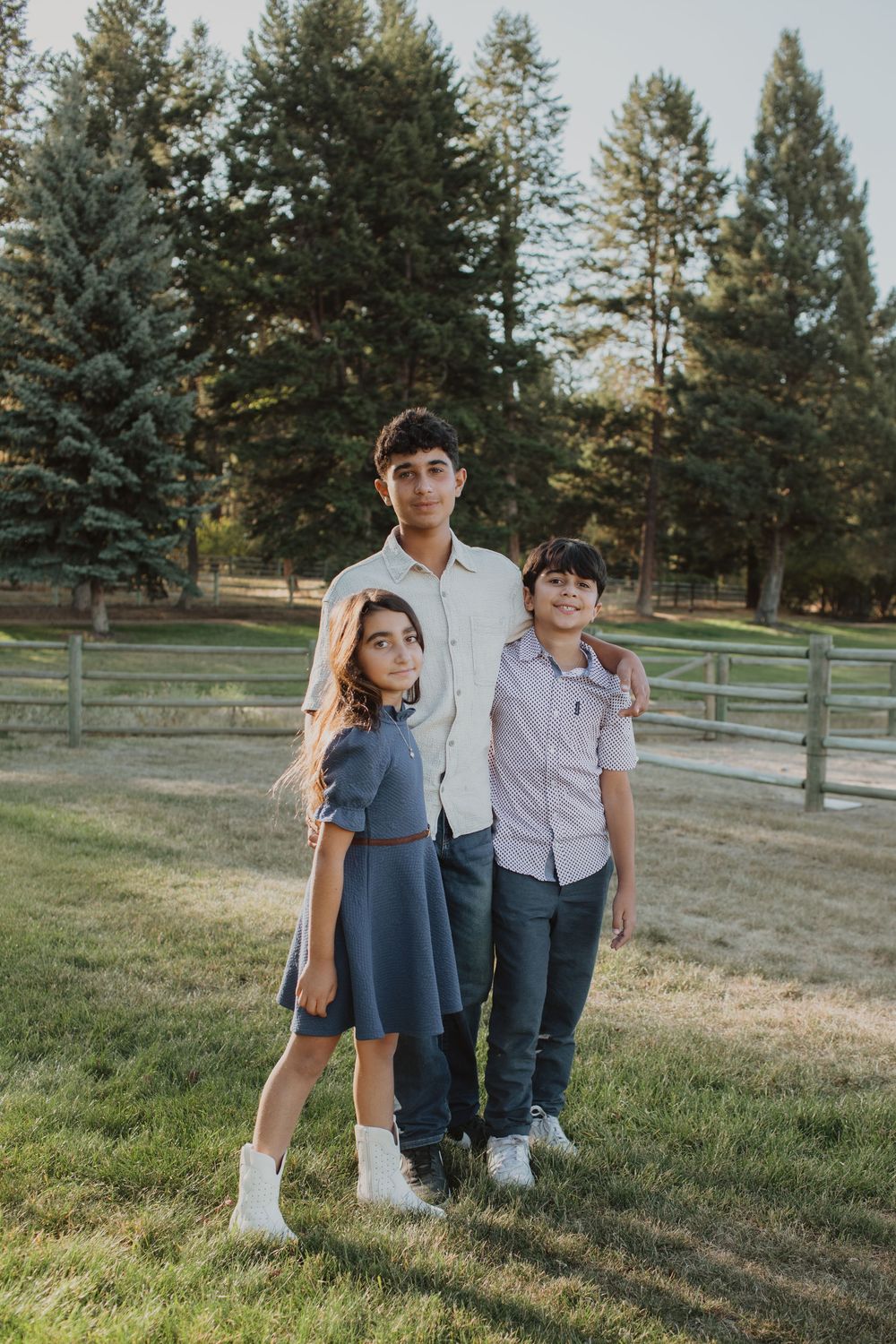 Three young people stand together in a grassy field with evergreen trees behind them.
