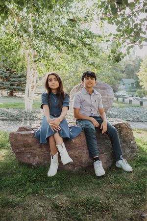 Two young people sit casually on a large rock under leafy trees.