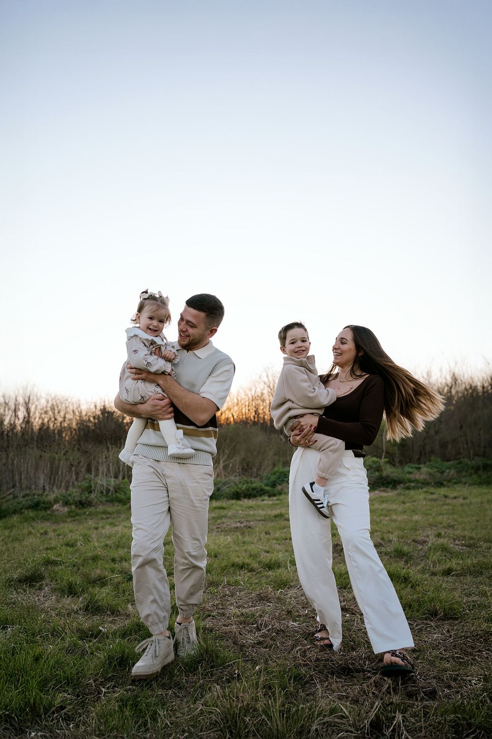 A family in coordinating cream-colored outfits plays and embraces together in a grassy outdoor setting at sunset.