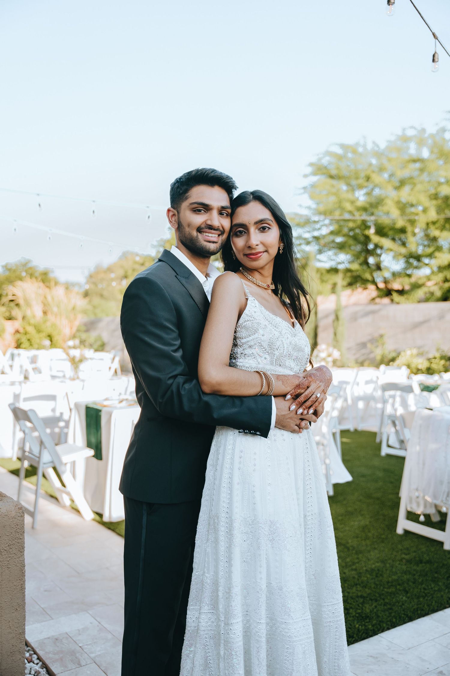 Couple embraces during outdoor wedding celebration with white chairs in background.