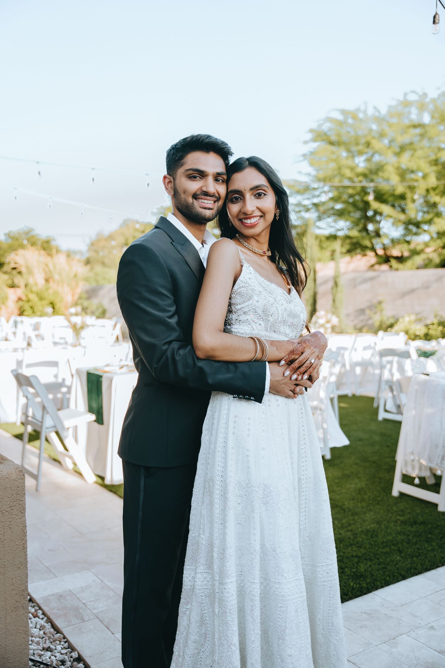A couple in wedding attire embraces at an outdoor venue with white chairs in the background.