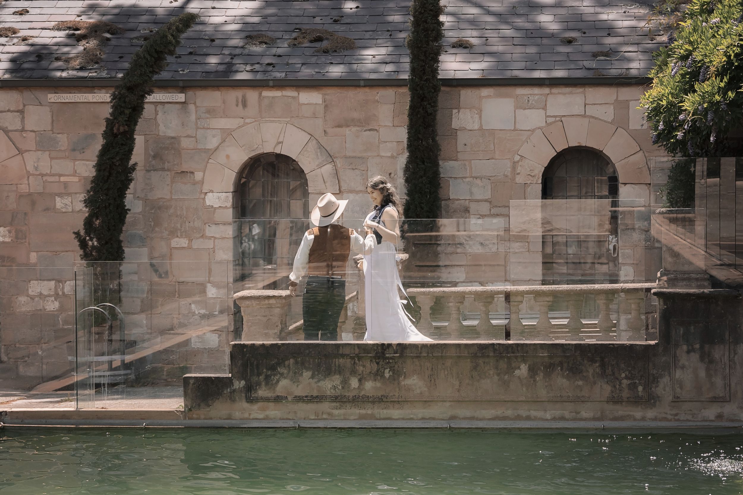 A romantic couple in wedding attire stands on a stone terrace overlooking a pond with cypress trees and arched walls.