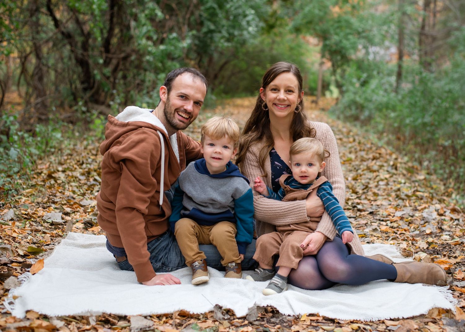 Family enjoying a fall day outdoors on a blanket surrounded by autumn leaves in a wooded setting.
