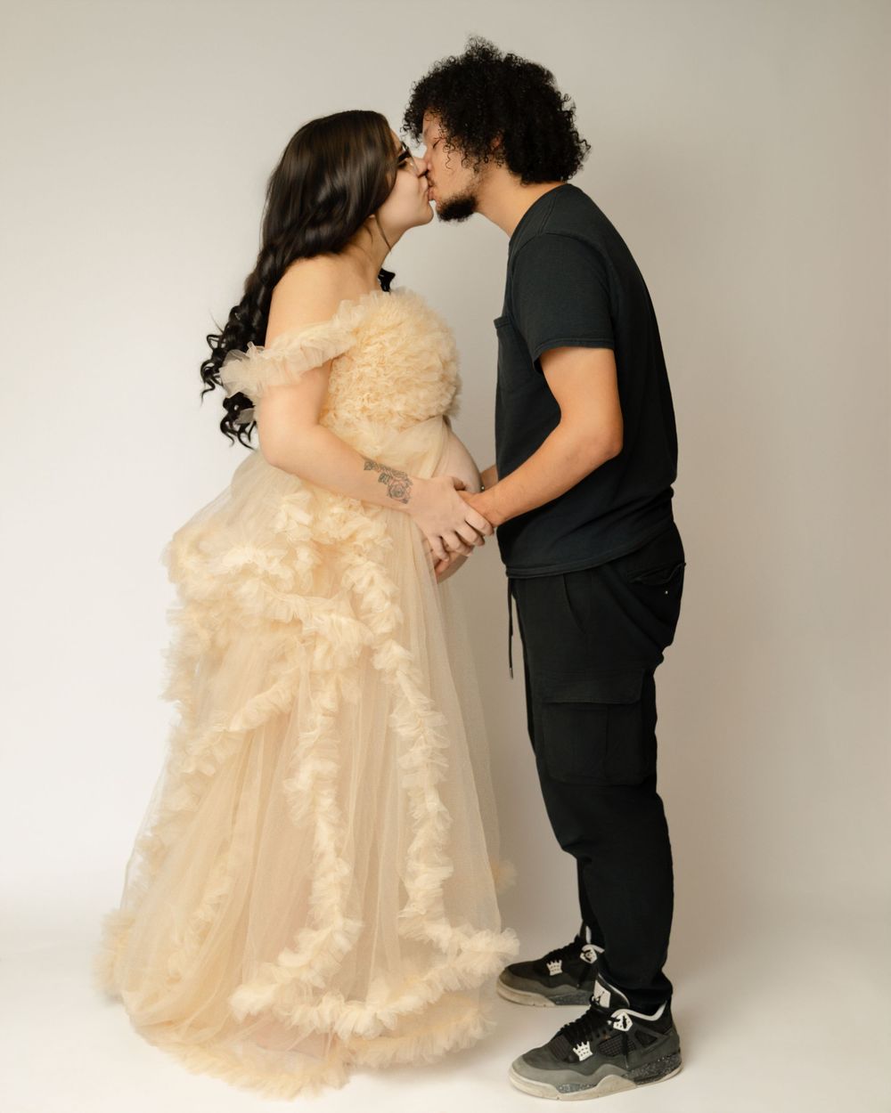 A romantic couple sharing a kiss, with one wearing a cream lace gown and the other in casual black attire against a neutral backdrop.
