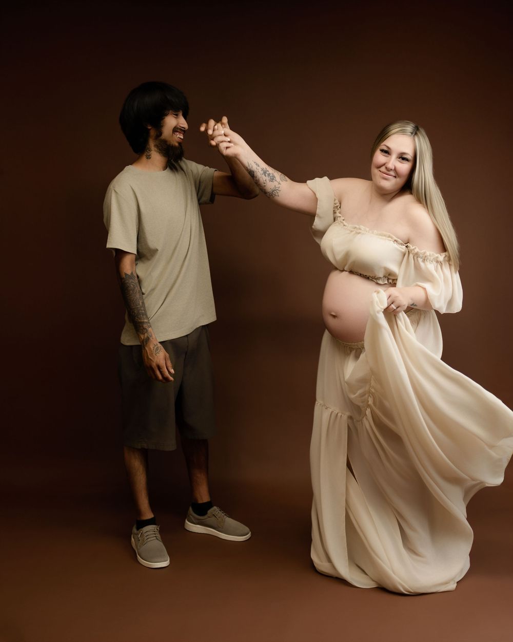 Mother in a white dress poses playfully with a child during studio maternity photography session.