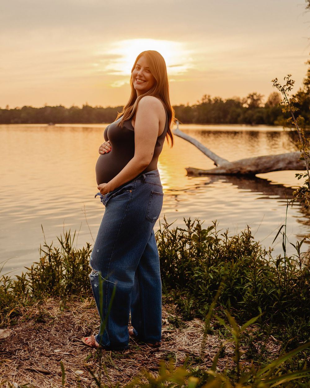 Maternity silhouette photo shoot at sunset by a lake with warm golden light filtering through the trees.