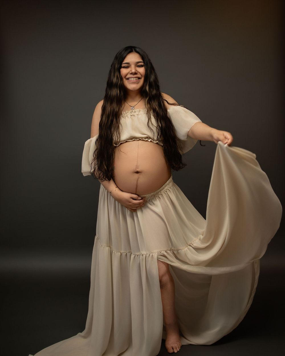 Elegant maternity photoshoot with figure draped in flowing beige fabric against dark background.
