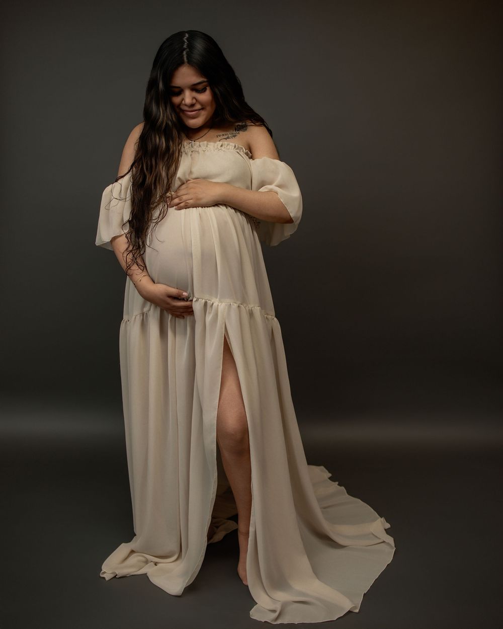 A person in a flowing white maternity gown poses gracefully against a dark background in this ethereal portrait series.