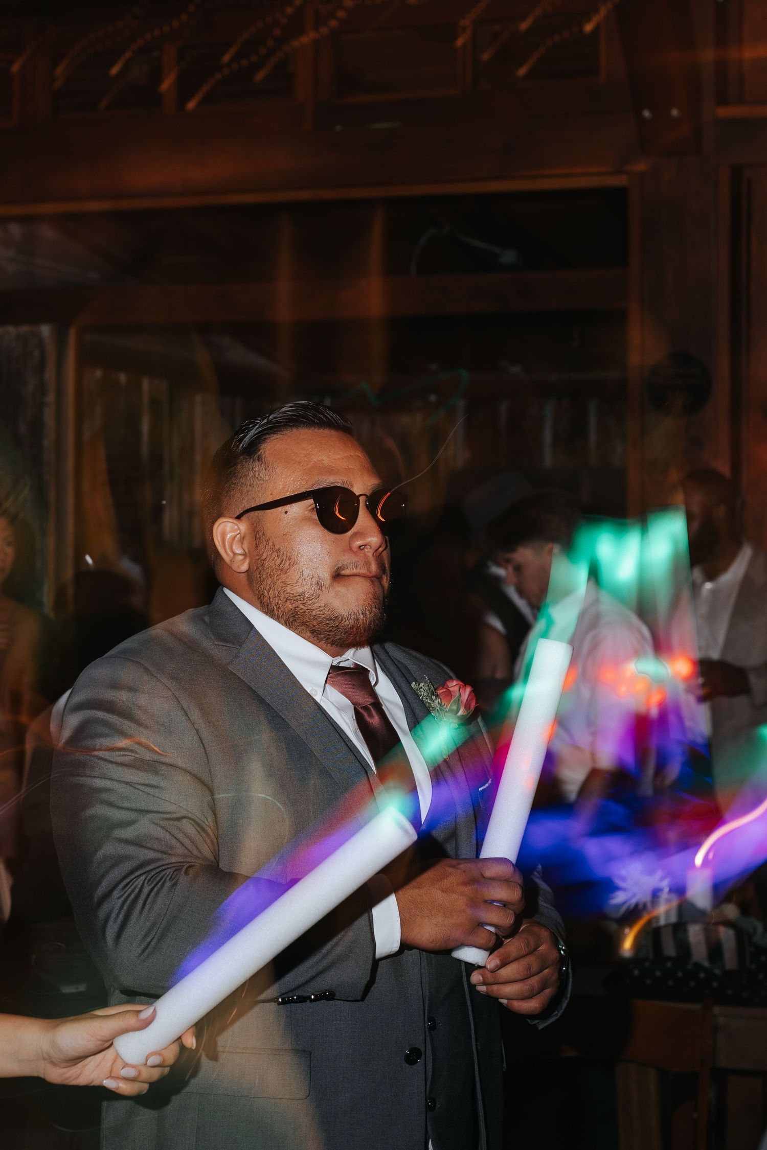 A guest in a gray suit waves glowing LED foam sticks at a wedding reception, creating colorful light trails. Knoxville Photographer