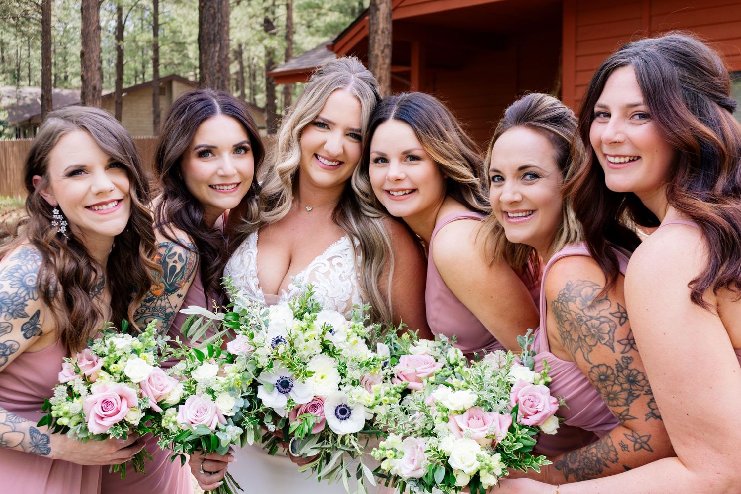 A group in soft mauve dresses stand together in an outdoor wooded setting holding white and green floral bouquets.