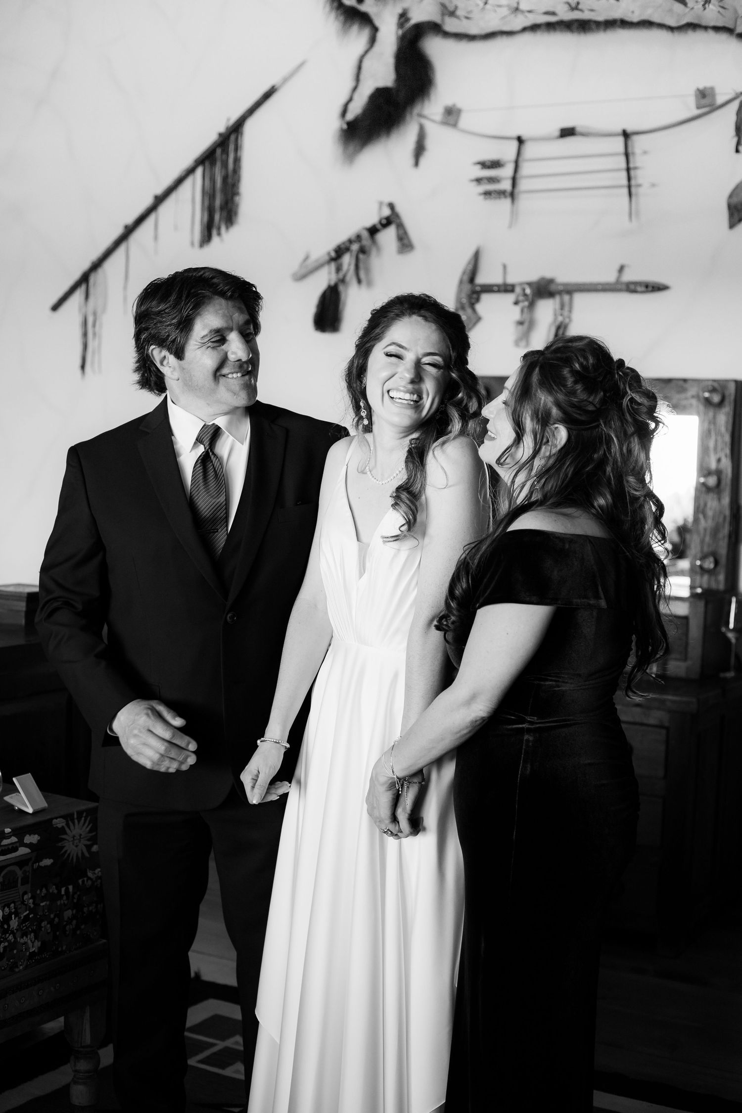 Black and white photo showing an intimate wedding moment with two women and a man sharing laughter indoors.
