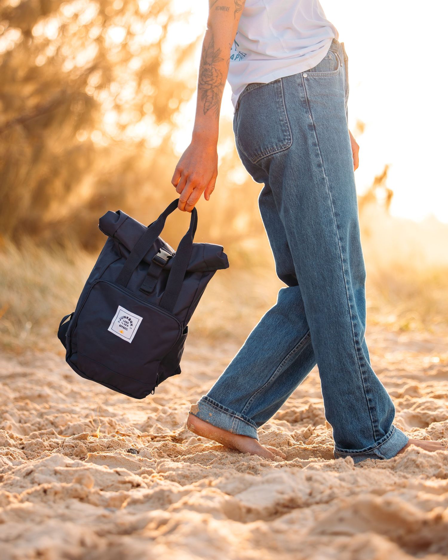 Person walking on sandy beach with navy blue backpack at golden hour.