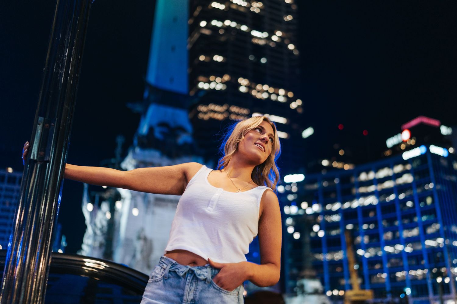 Woman in casual attire poses against cityscape with illuminated skyscrapers at night.