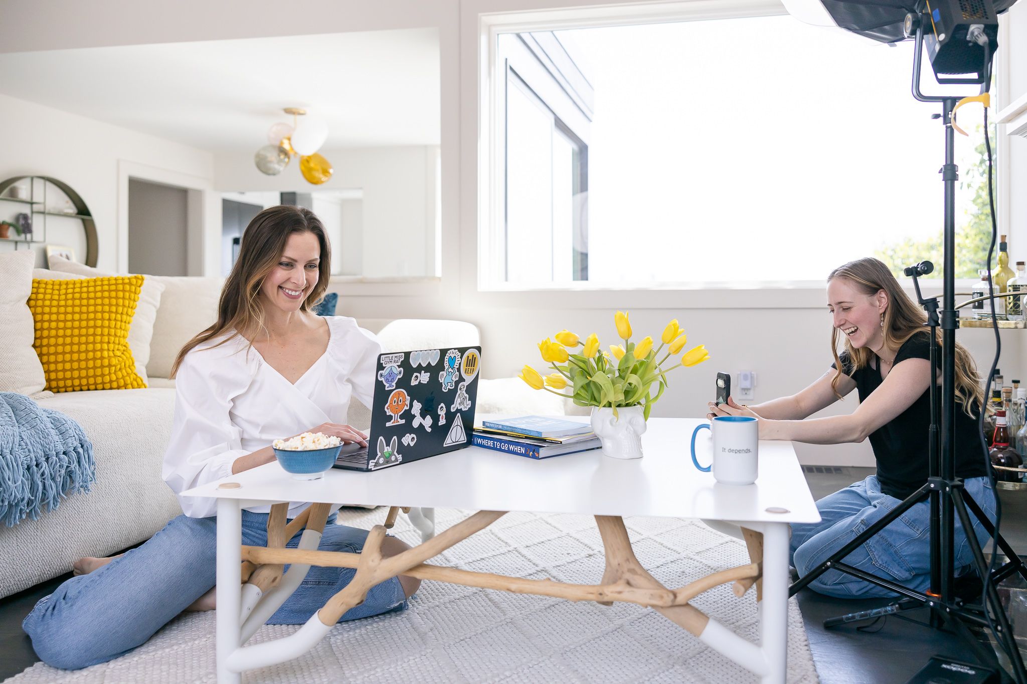 two women sitting on floor for behind the scenes branding photoshoot in Seattle, WA home 1 woman on computer while other films on phone