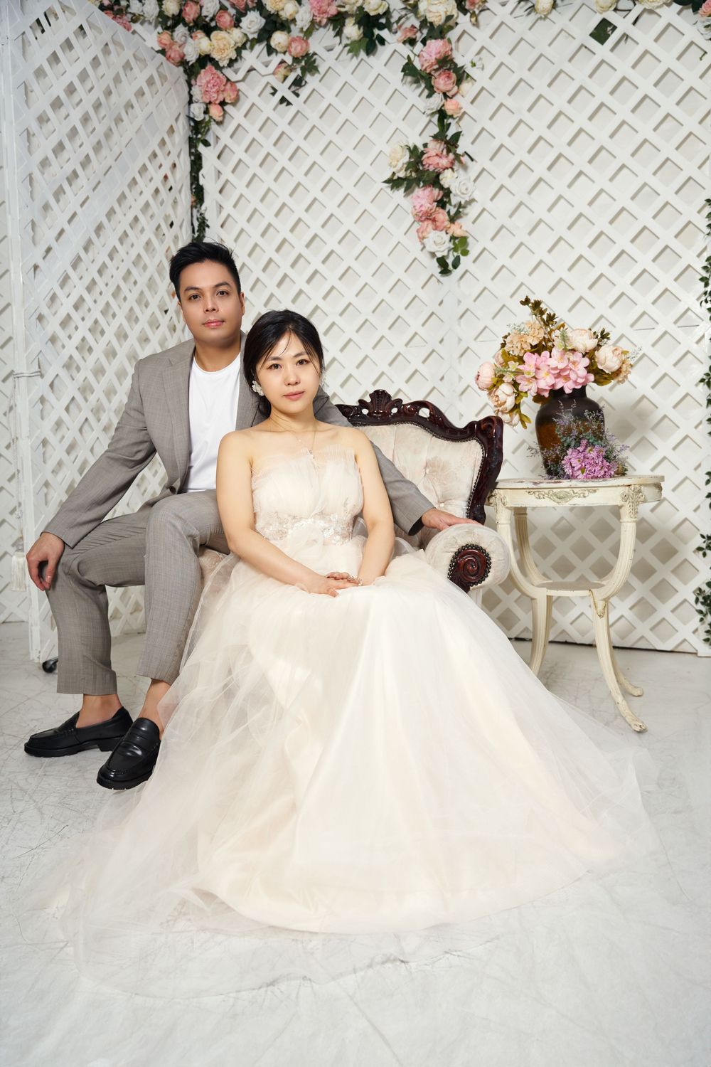 Newlywed couple poses together against white lattice backdrop decorated with pink and cream floral arrangements. studio photo session