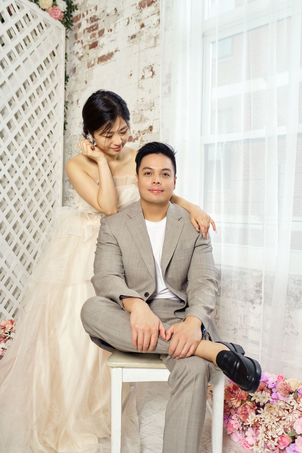 Couple in wedding attire poses together near white lattice wall and pink floral decorations in soft natural light. studio photo session