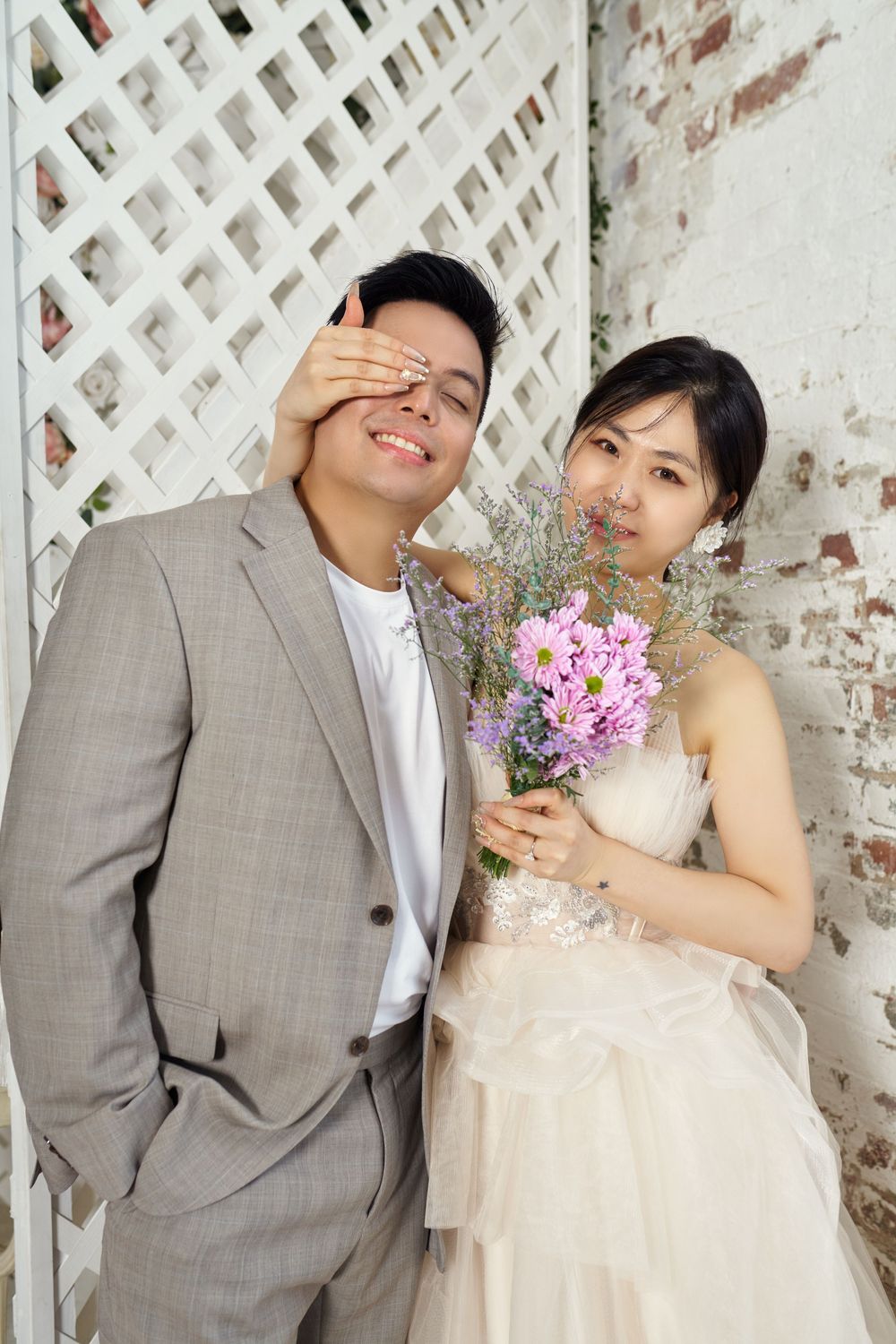 A playful wedding portrait with the couple posing against a white lattice and brick wall, holding a pink floral bouquet. studio photo sessio