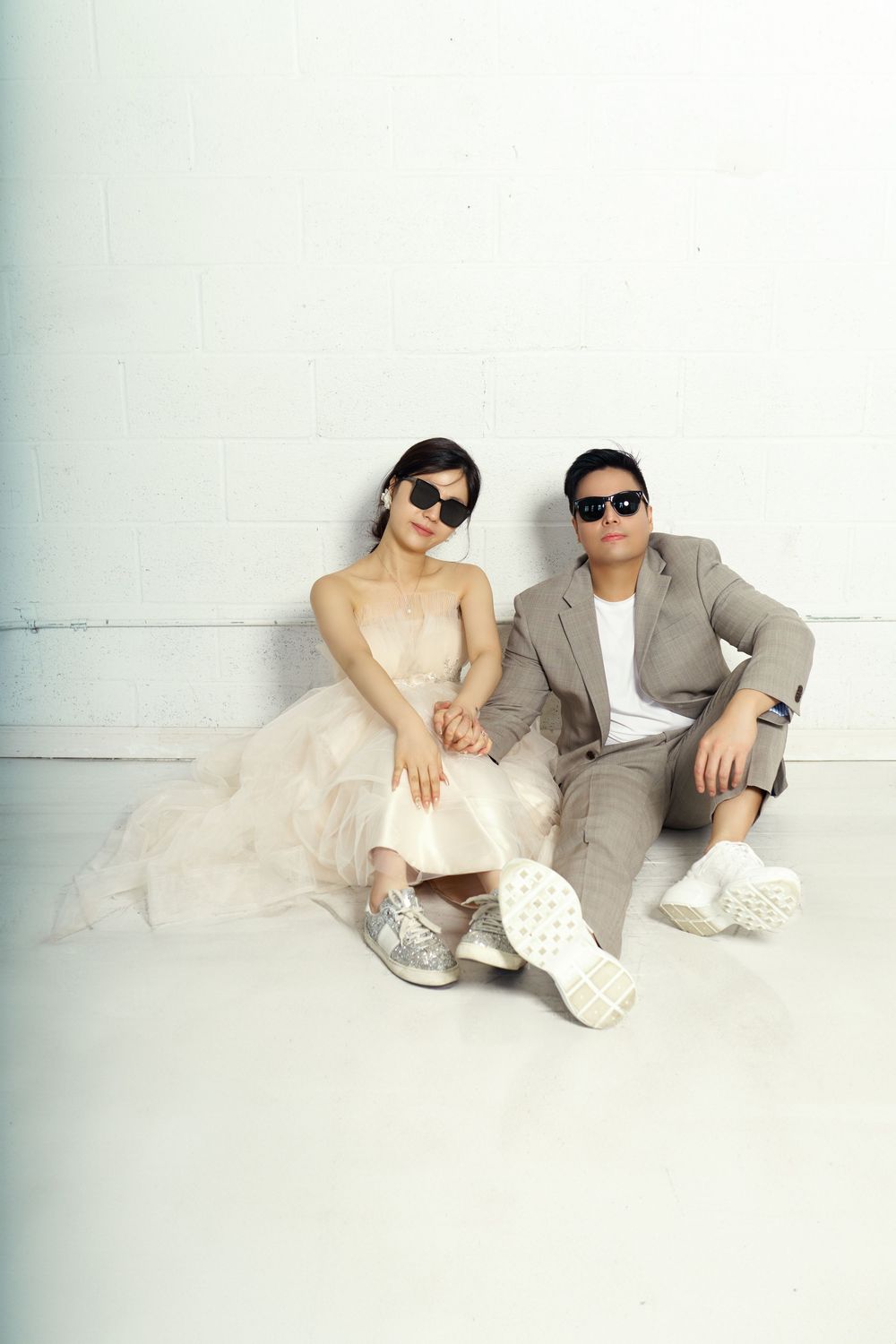 A cool modern couple in sunglasses and casual attire lounges against a white wall wearing matching sneakers. studio photo session