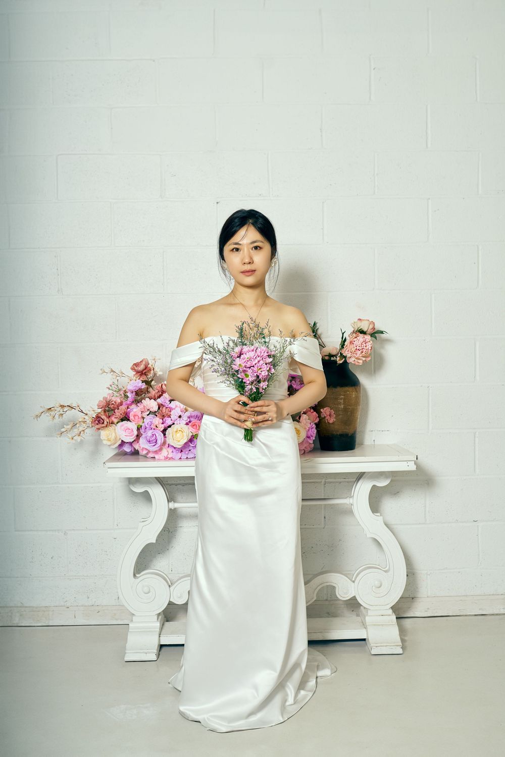 An elegant bridal session featuring a white satin wedding dress, white ornate table, and pink floral arrangements, studio photo session