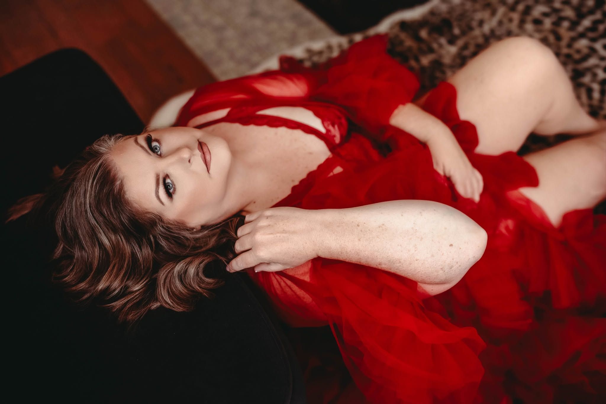 Woman with short auburn hair lying back in a flowing red tulle dress during a boudoir photography session with soft, warm lighting.