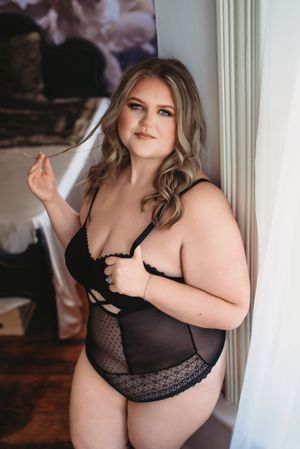 Woman with wavy blonde hair in black lace bodysuit poses by a sheer curtain, twirling a strand of hair in a boudoir portrait.