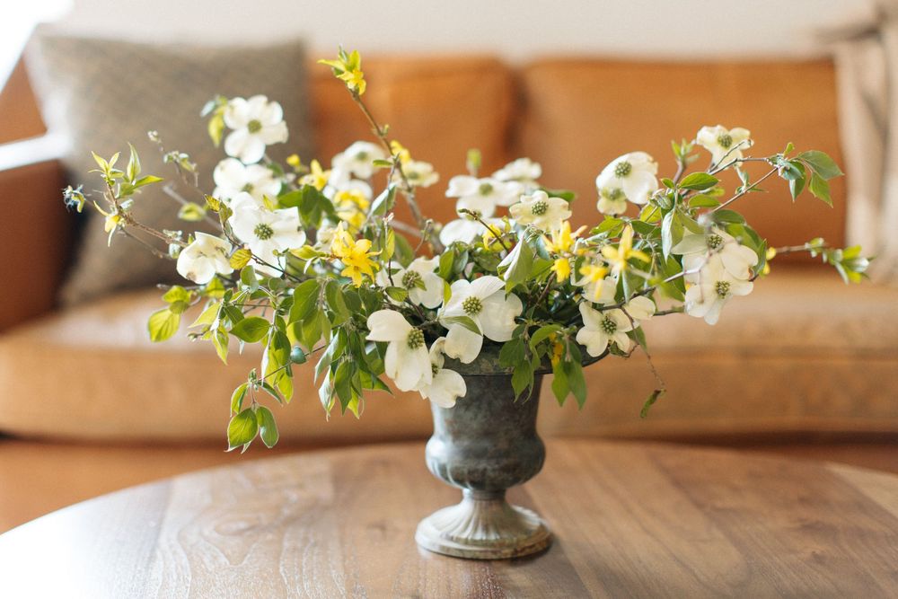 White and yellow spring flowers arranged in a gray stone urn vase on wooden table with tan sofa in background.