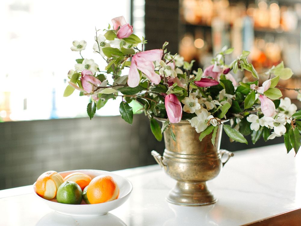 Beautiful pink and white floral arrangement in brass vase with citrus fruits in bowl on white marble table.