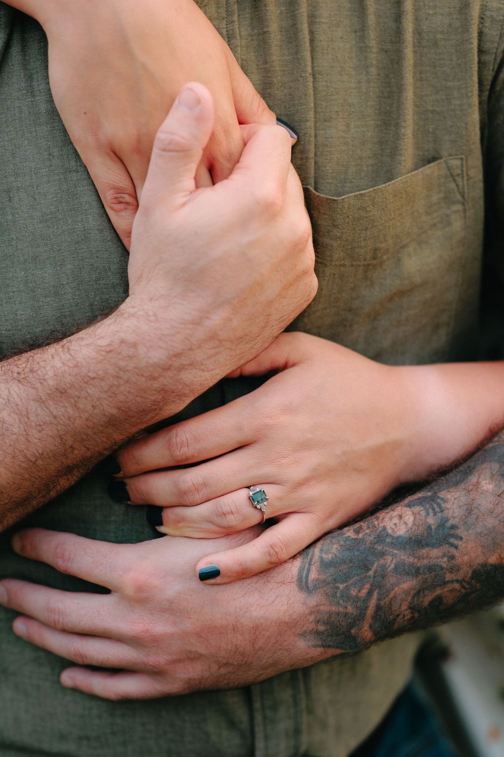 Close-up of hands embracing with a diamond ring and visible tattoo against an olive green background.