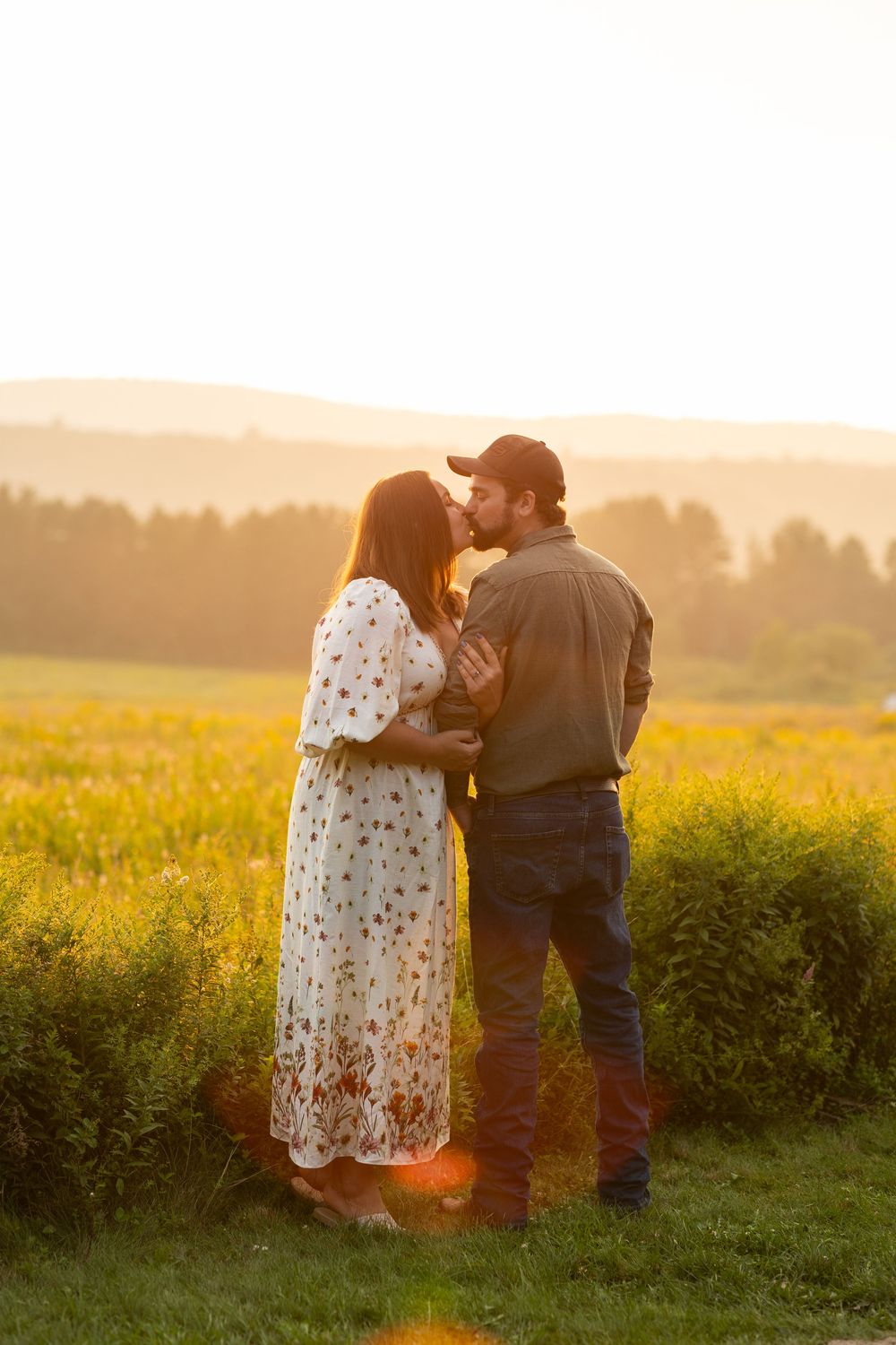 A romantic couple embraces in a golden meadow at sunset while surrounded by wildflowers and rolling hills.