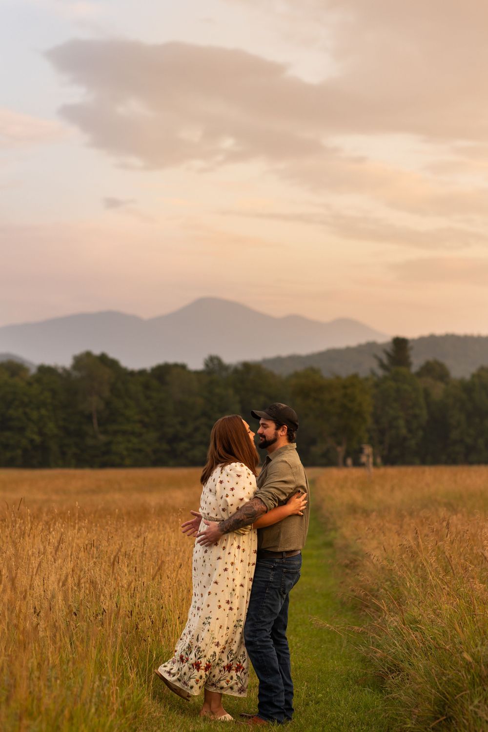 A couple stands together in a grassy meadow at sunset with mountains and trees visible in the distance.