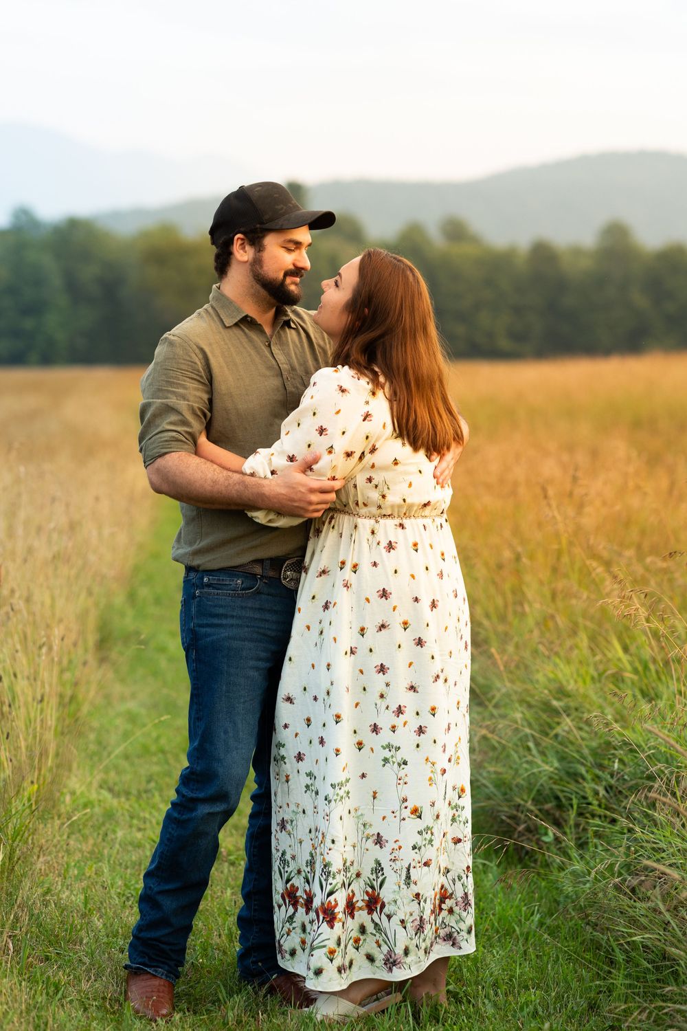 A romantic couple embraces in a golden meadow with mountains in the background at sunset.