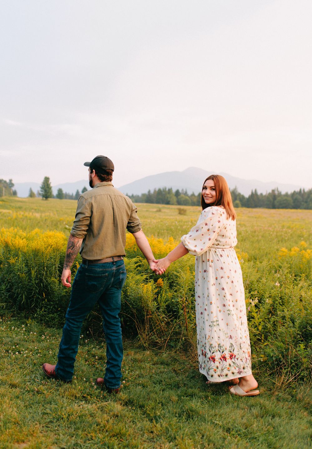 A couple walks through a flowering meadow at sunset holding hands with mountains visible in the distance.