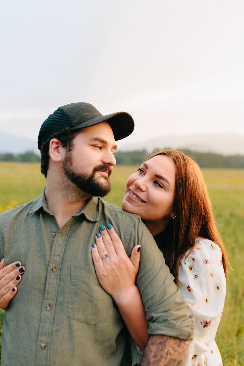Couple smiles lovingly at each other while embracing in a sunny green field at sunset.