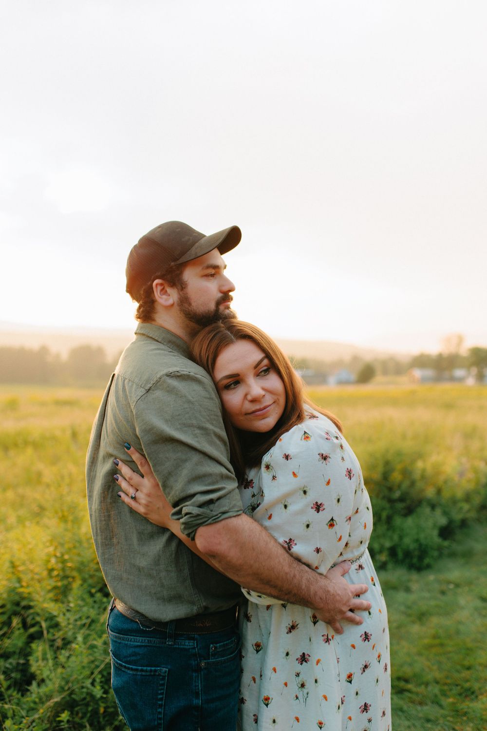 A couple embraces in a field at sunset, with the person wearing a baseball cap and green shirt hugging their partner.