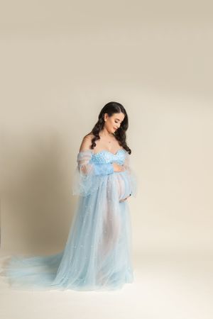 Pregnant woman wearing ethereal powder blue tulle gown with off-shoulder design in dreamy maternity photoshoot.