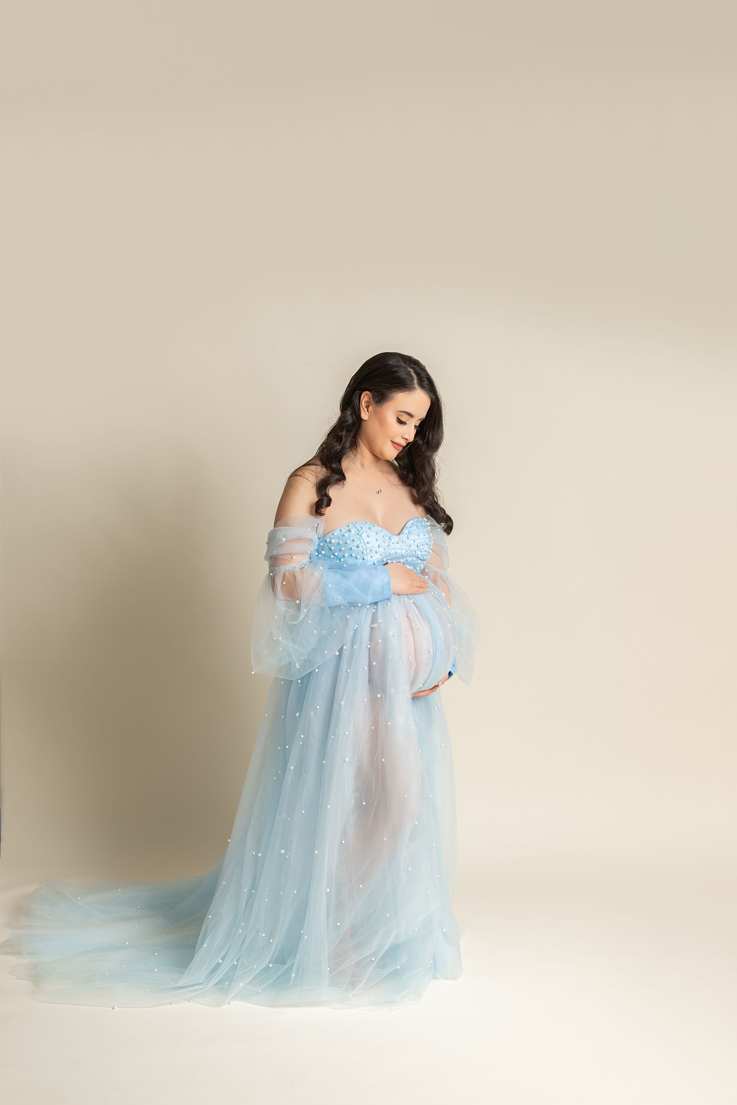 Pregnant woman wearing ethereal powder blue tulle gown with off-shoulder design in dreamy maternity photoshoot.