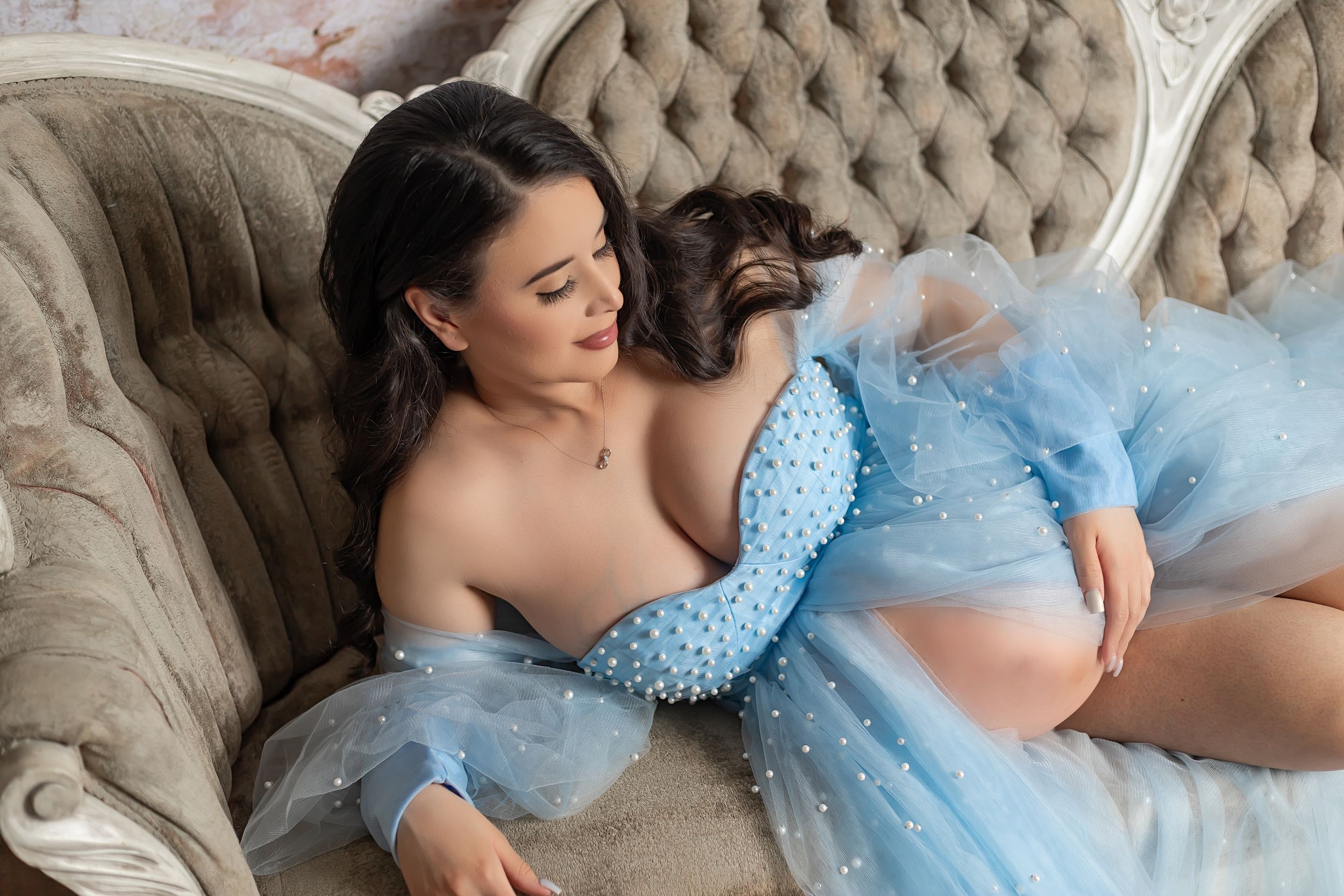 Expectant mother in blue tulle robe reclining on vintage beige tufted sofa during maternity photoshoot.