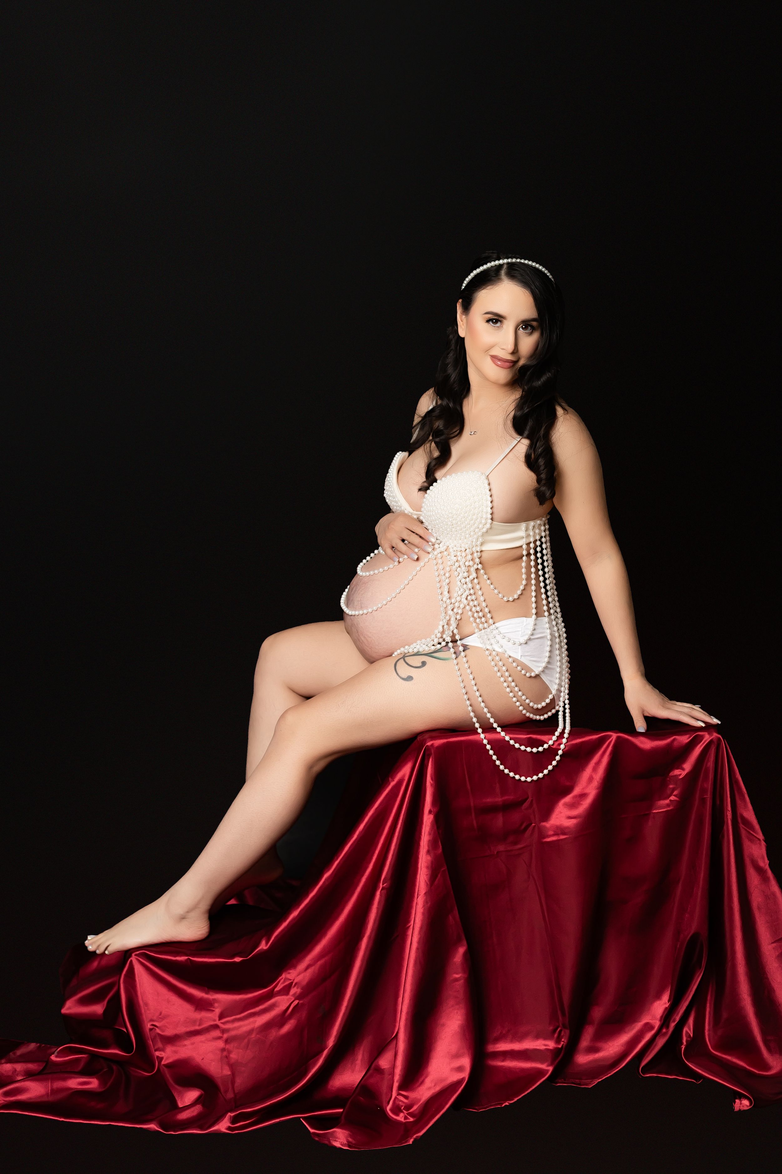 An elegant maternity portrait with a figure in white pearls reclining on luxurious red satin fabric against a dark backdrop.