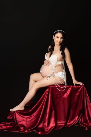 An elegant maternity portrait with a figure in white pearls reclining on luxurious red satin fabric against a dark backdrop.