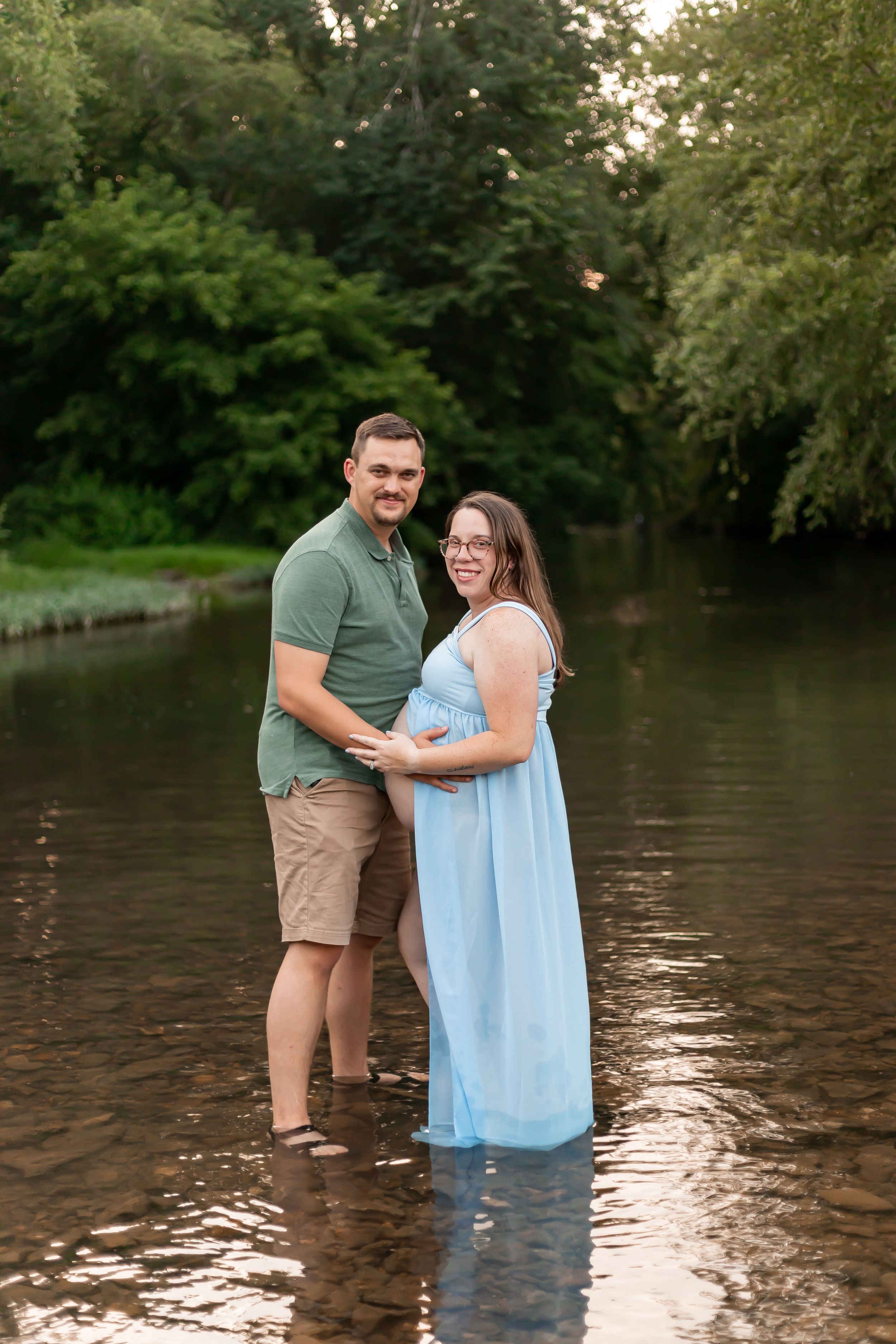 A couple stands together in shallow water surrounded by green trees on a summer evening.
