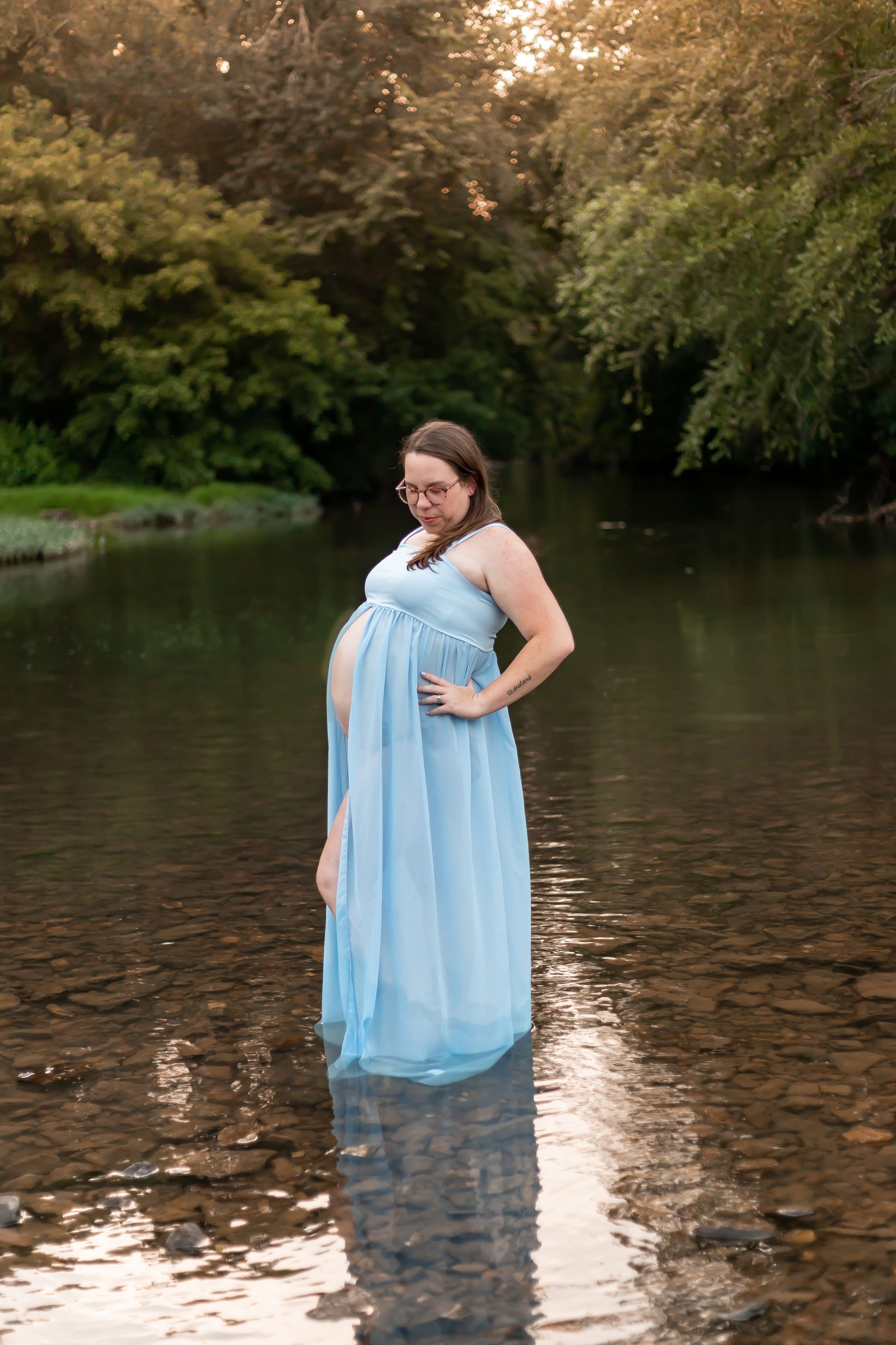 A figure in a flowing light blue dress stands in calm waters surrounded by lush greenery during golden hour.