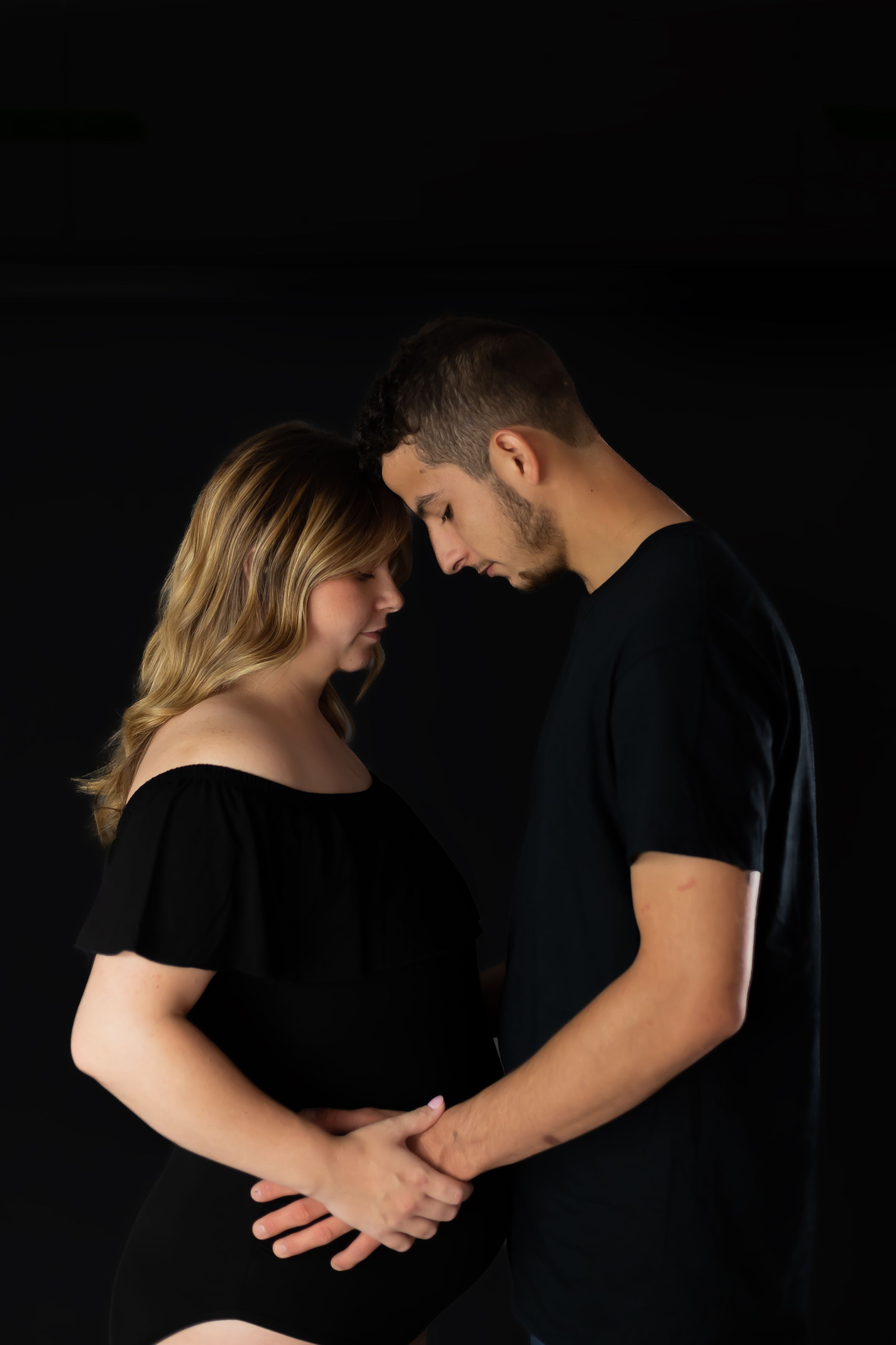 A couple in black attire shares an intimate moment against a dark backdrop, holding hands while standing close together.