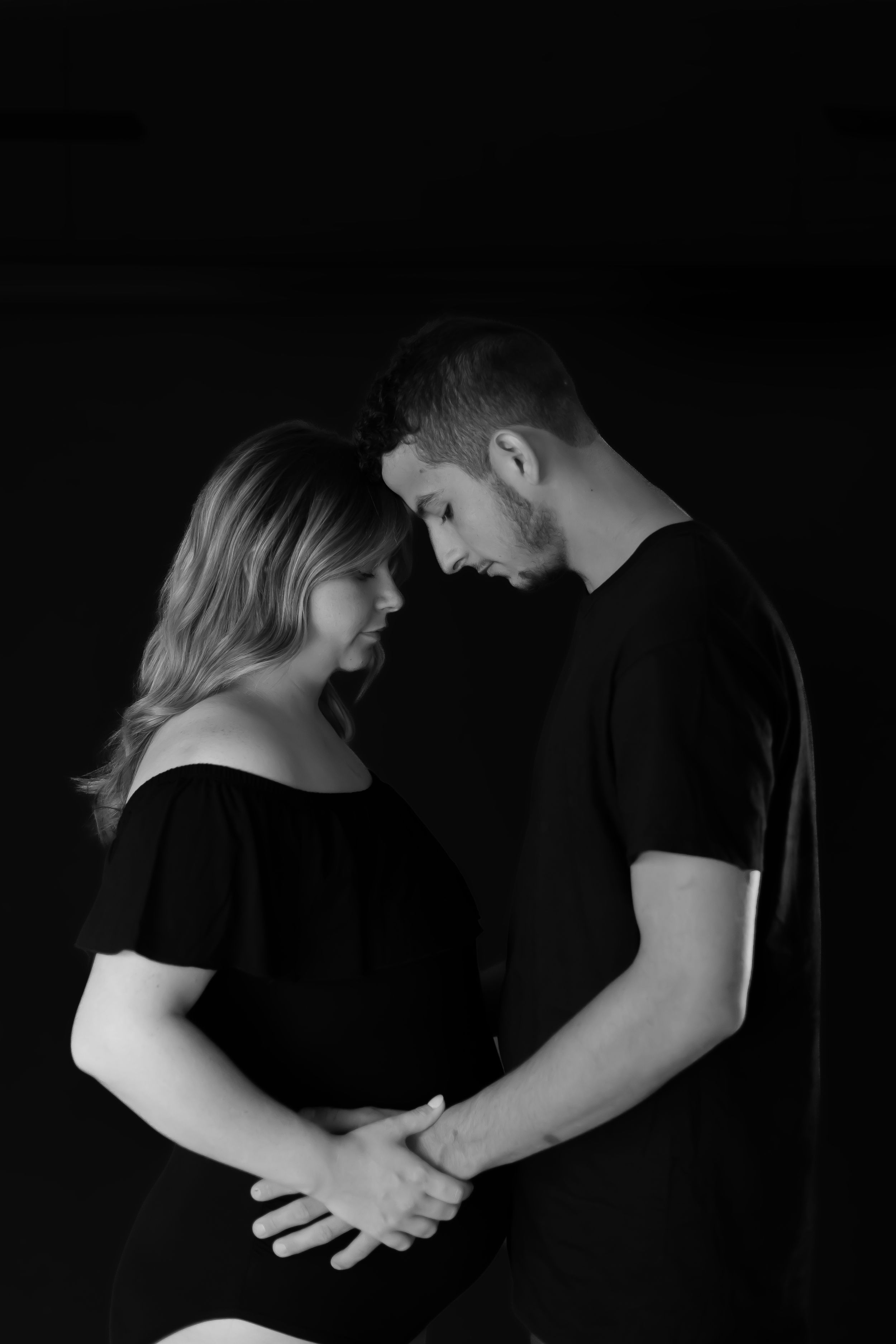 A tender black and white portrait of a couple holding hands against a dark background.