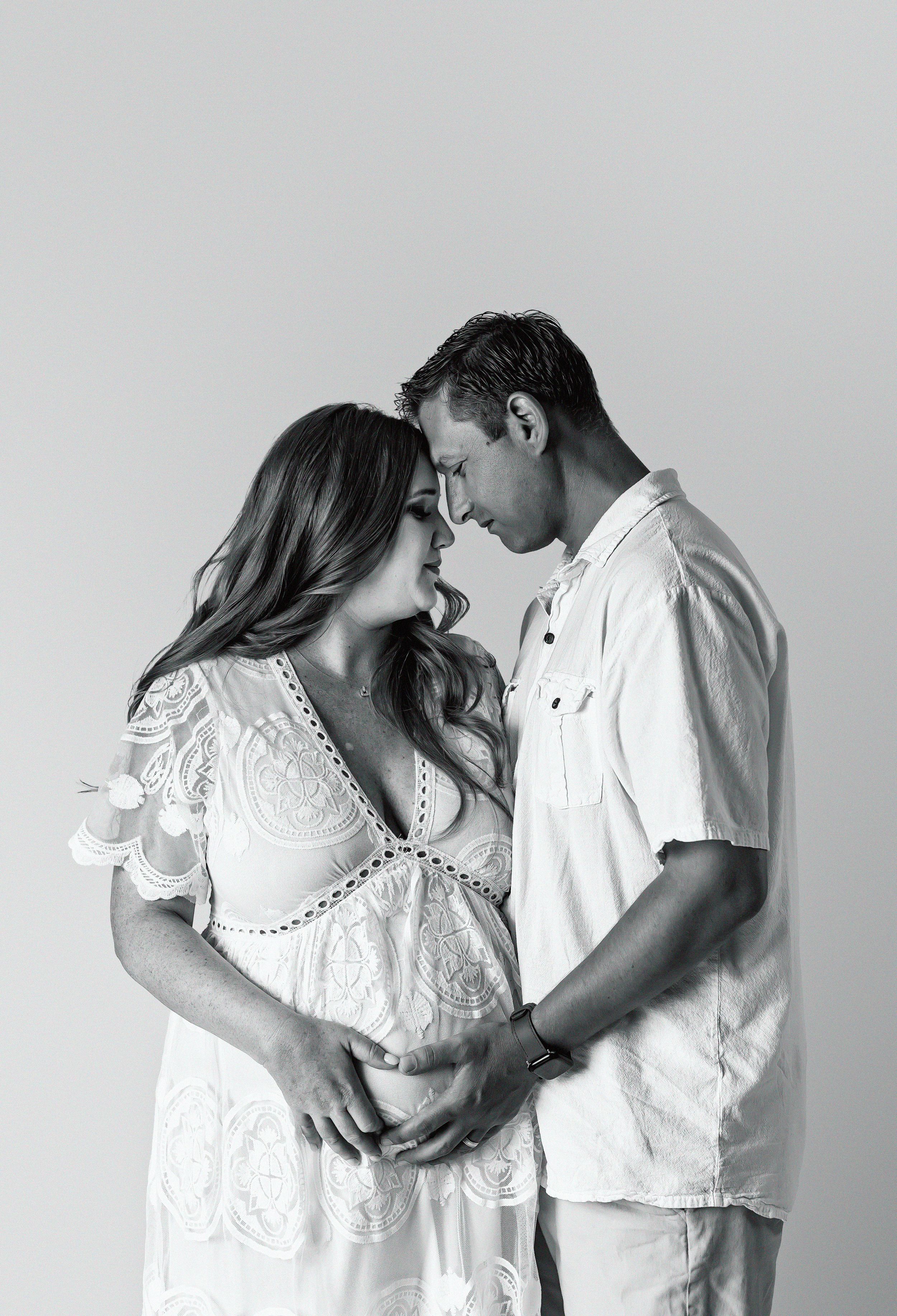 Intimate black and white maternity photo of couple in white clothing sharing a tender moment together.