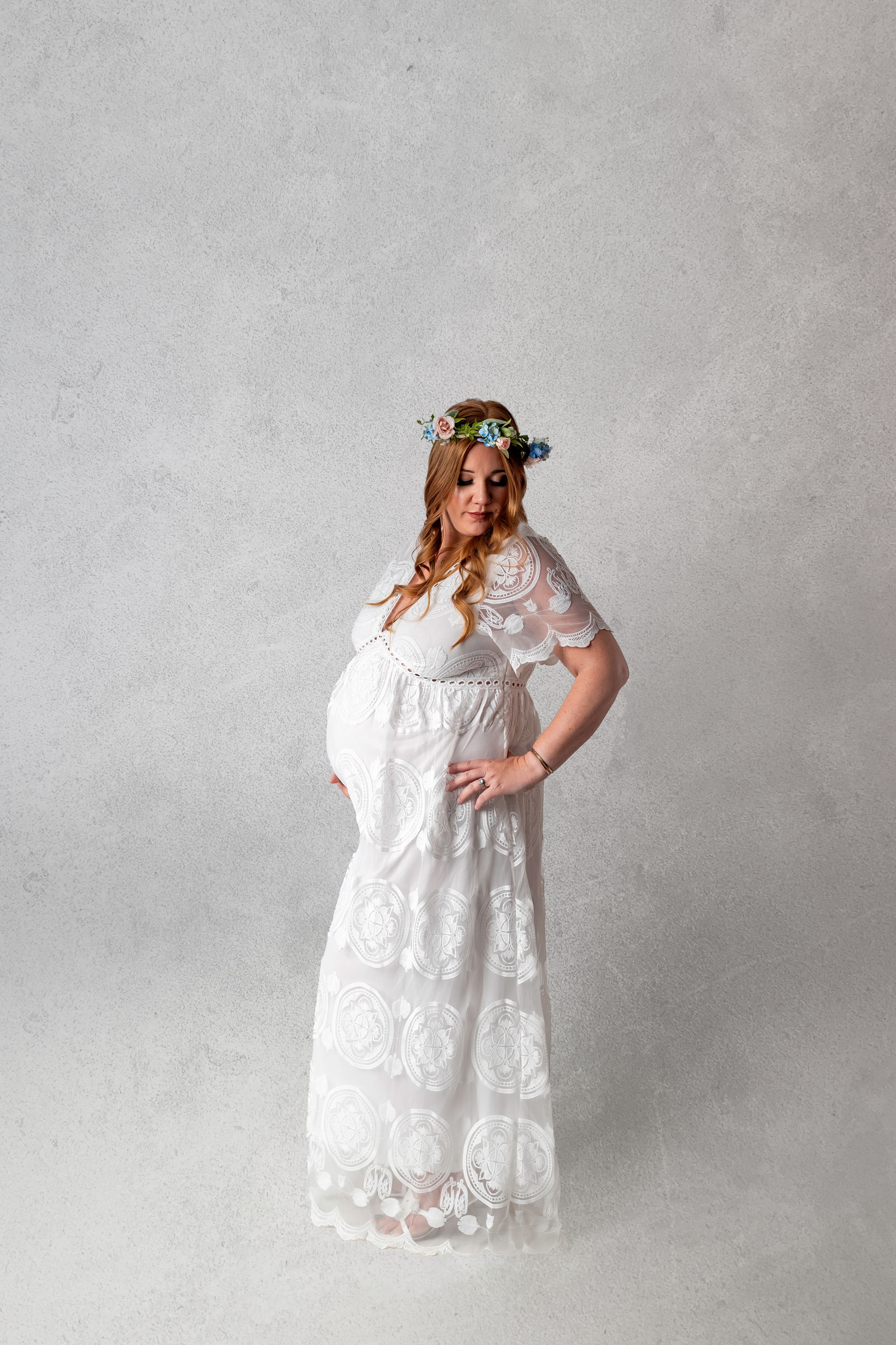 Ethereal maternity portrait featuring a flowing white lace maxi dress and delicate blue floral crown against grey backdrop.
