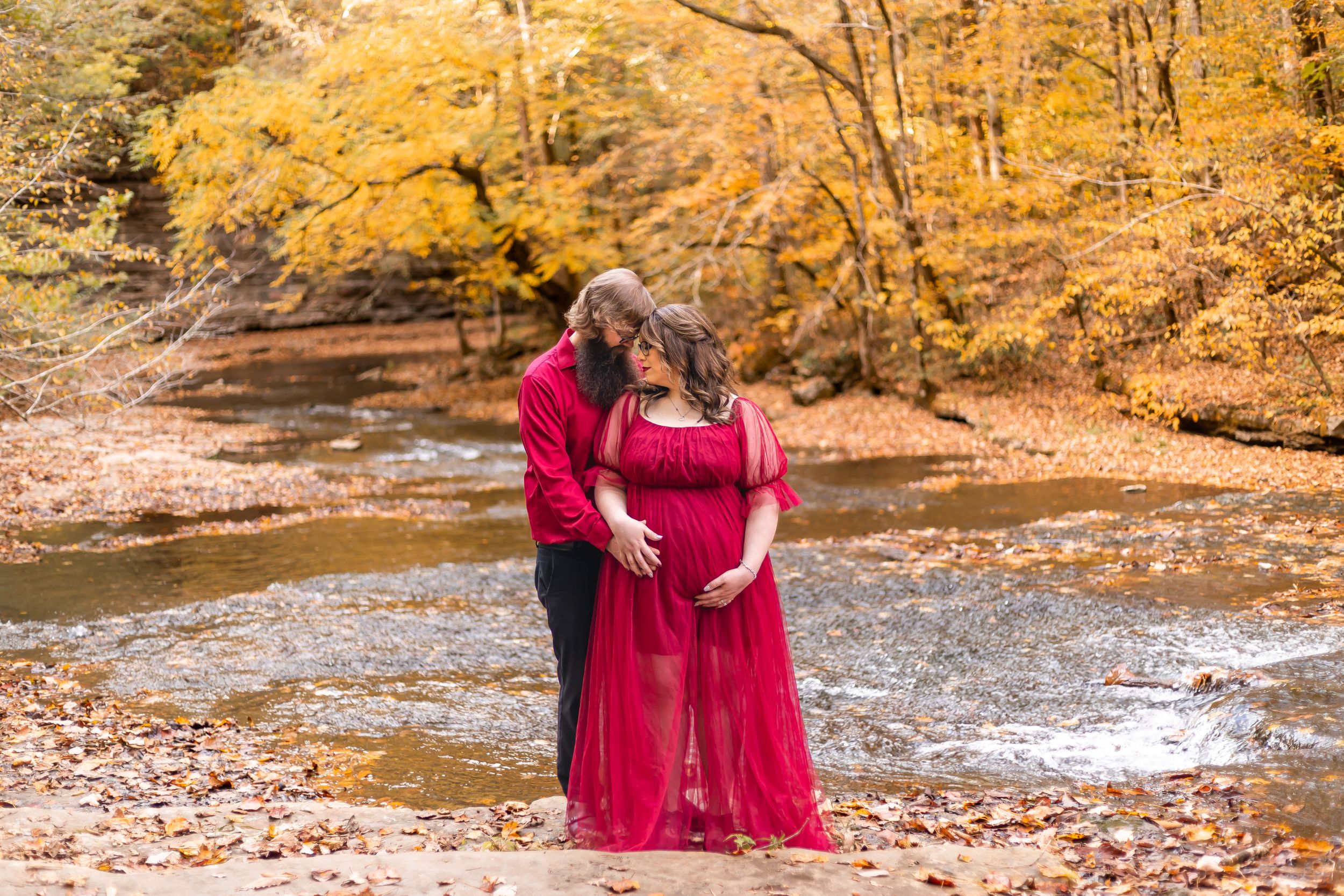A couple shares a tender moment by a creek surrounded by golden autumn foliage in a forest setting.