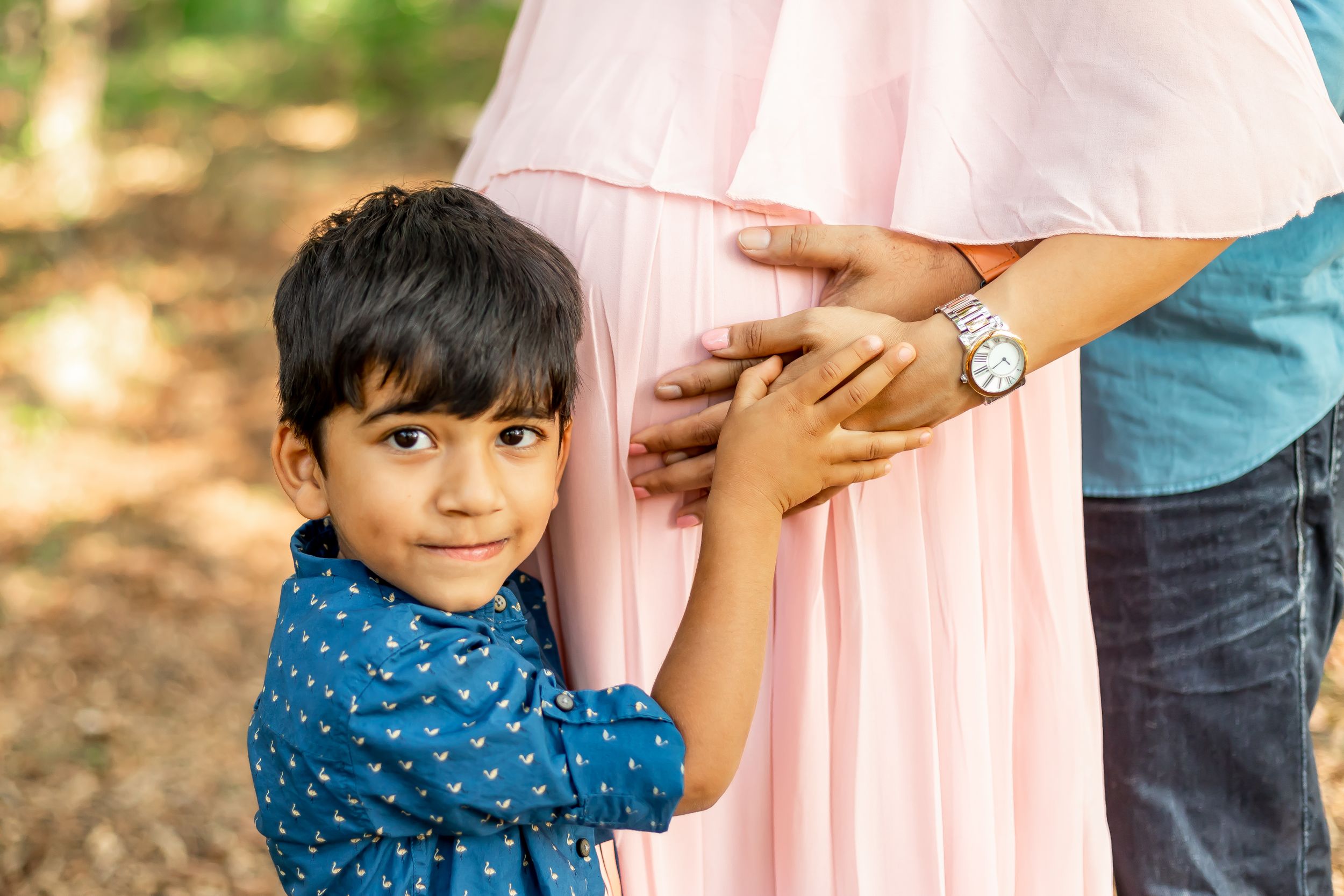 A young boy in a blue polka dot shirt hugs close to a person wearing a pink dress in an outdoor setting.